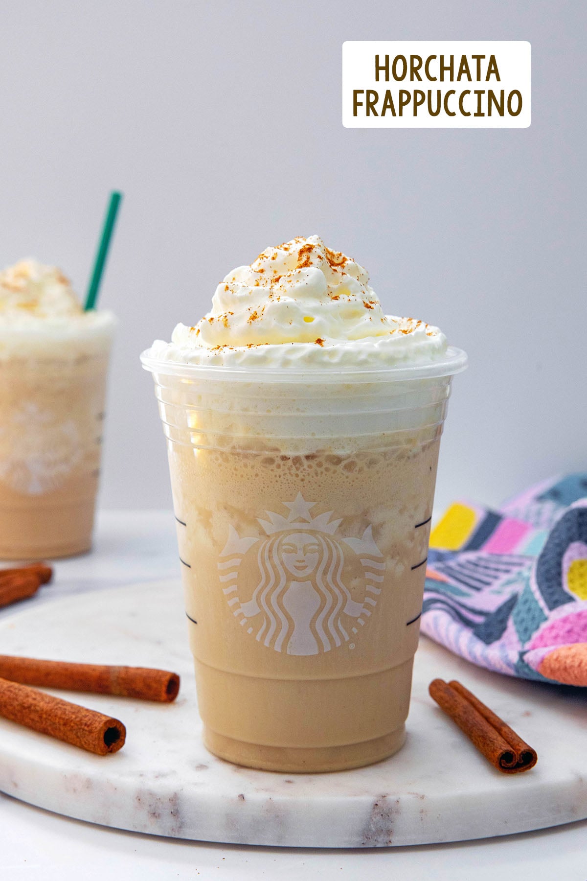 Head-on view of a Horchata Frappuccino in a Starbucks cup with cinnamon sticks around, second Frappuccino in background, and recipe title at top.