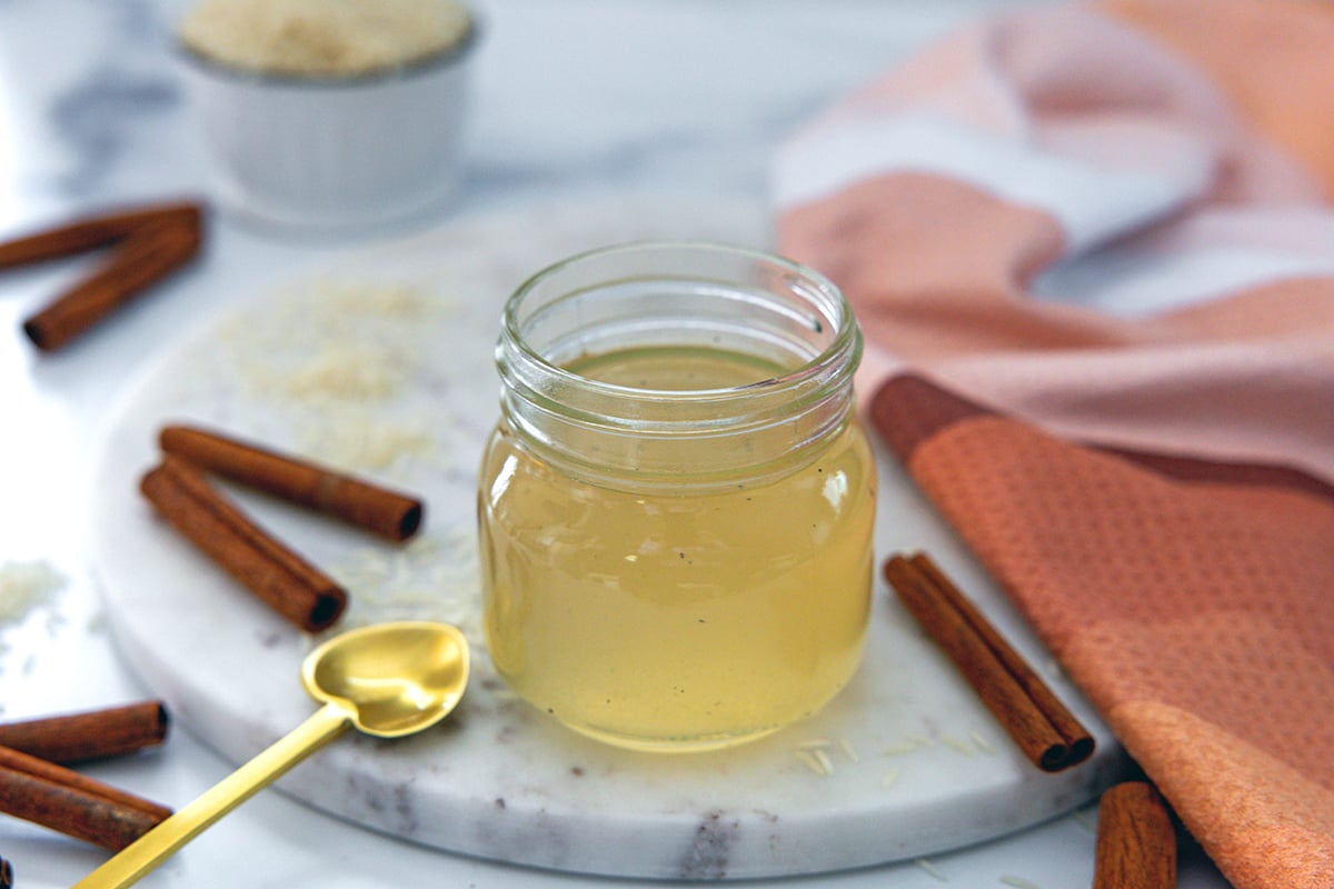 Small jar of horchata syrup with heart-shaped spoon next to it, along with cinnamon sticks and rice all around.
