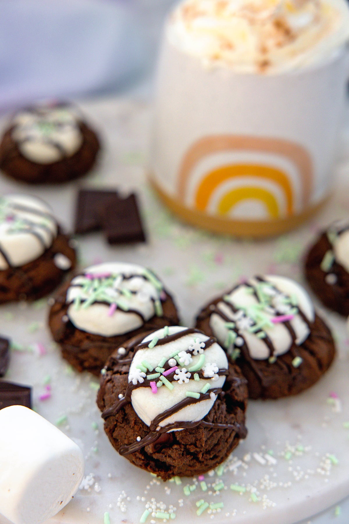 Hot chocolate cookies with marshmallows, chocolate drizzle, and sprinkles with a mug of hot chocolate in the background.