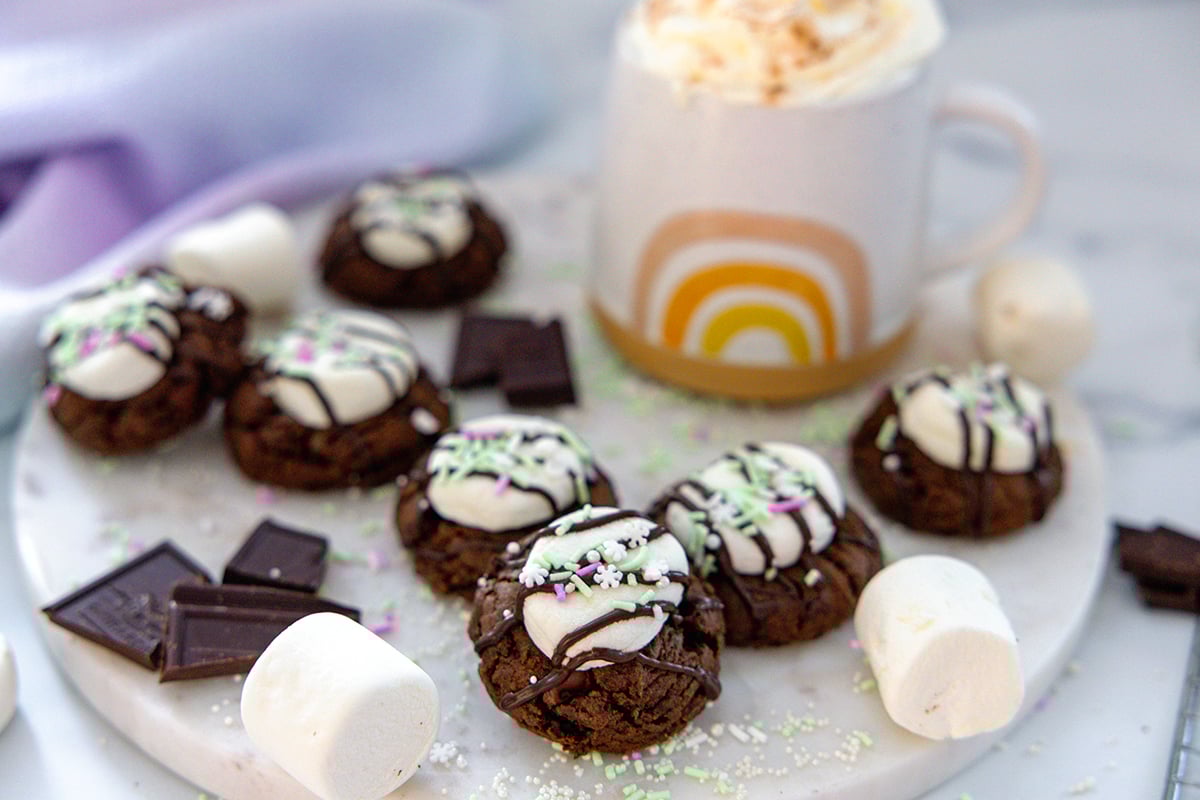 Landscape head-on view of hot chocolate cookies on a marble platter with marshmallows, chocolate pieces, and a mug of hot chocolate in background.