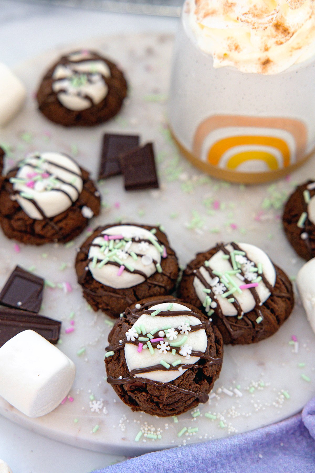 Overhead view of hot chocolate cookies with marshmallows and chocolate all around and mug of hot chocolate in background.