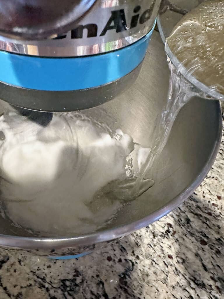 Hot corn syrup mixture being poured into egg whites in stand mixer.