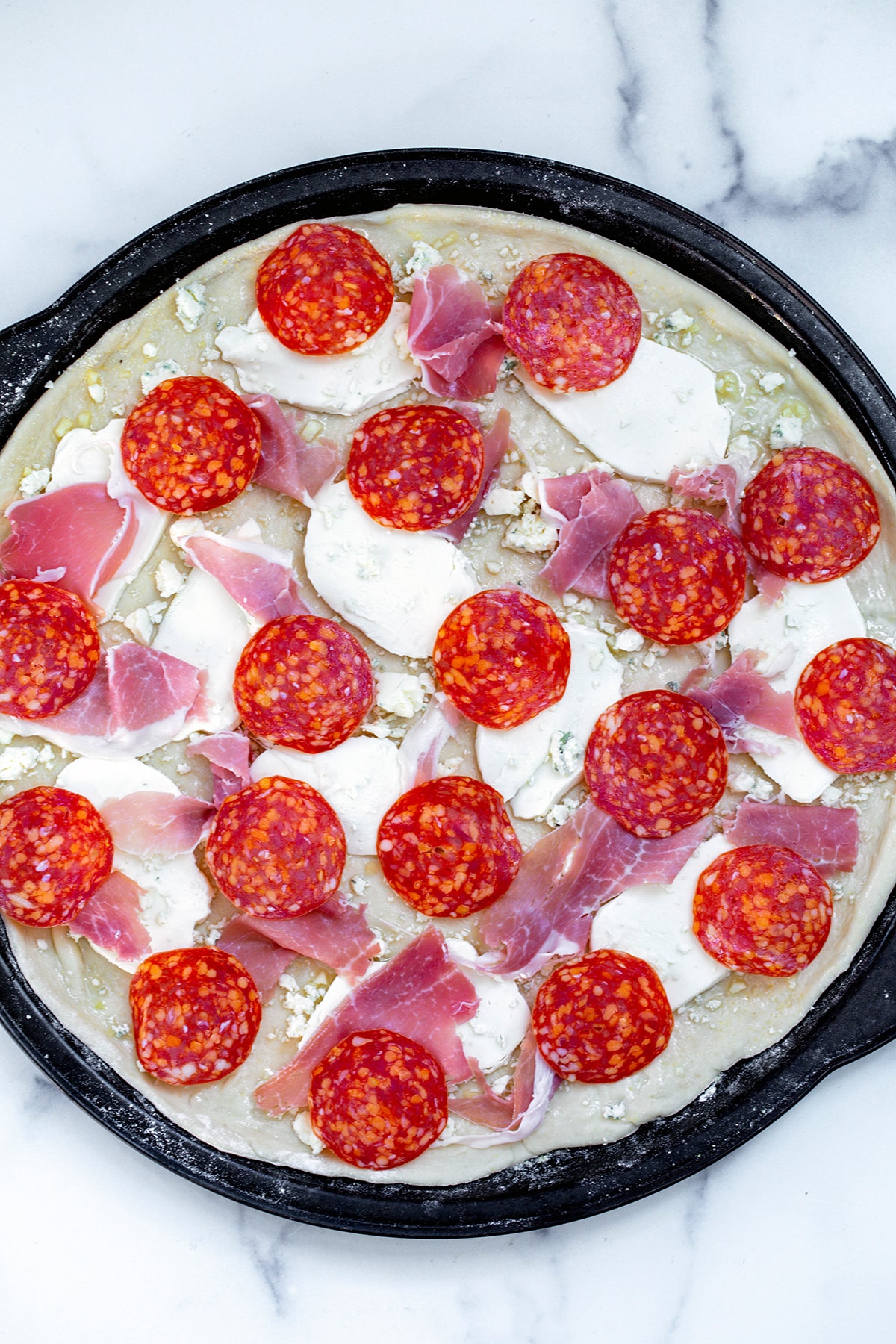 Pizza dough rolled out on round pan with sliced mozzarella, crumbled gorgonzola, spicy capicola, and prosciutto.