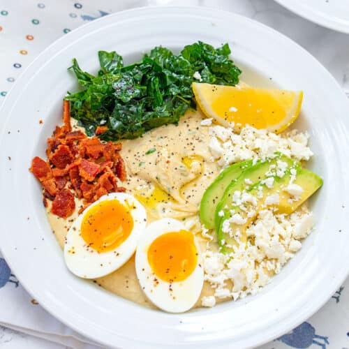 Closeup view of a hummus breakfast bowl with hardboiled egg, bacon, avocado, kale, feta, and lemon.