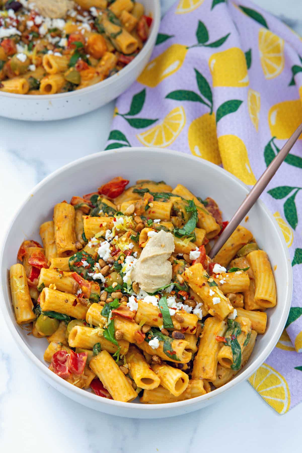Hummus pasta in a bowl with tomatoes, basil, olives, and scoop of hummus on top.