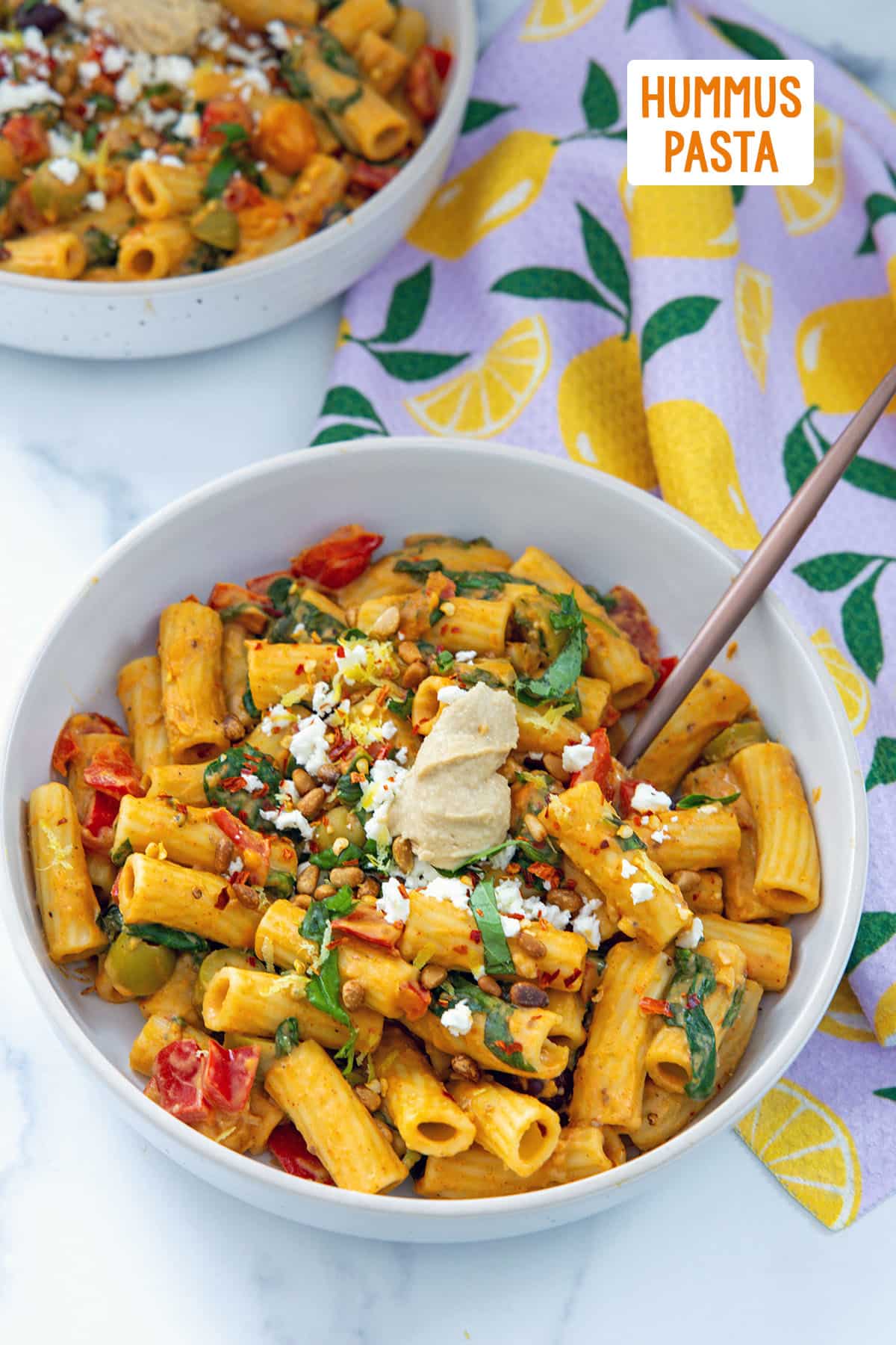 Hummus pasta in a bowl with tomatoes, basil, olives, and scoop of hummus on top with recipe title on top of image.