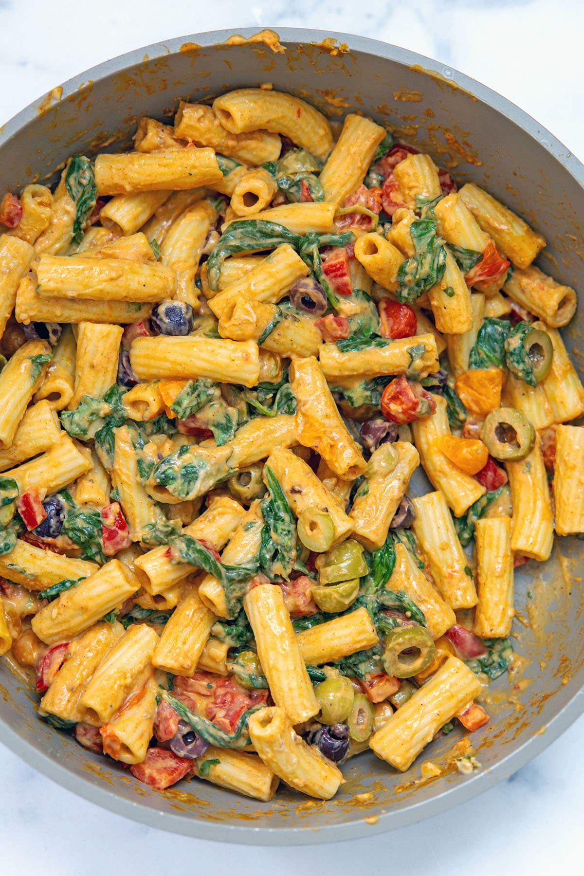 Hummus pasta with spinach, tomatoes, peppers, and olives tossed together in sauce in skillet.
