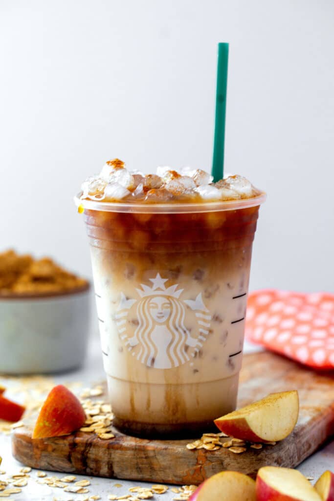 Head-on view of an Iced Apple Crisp Oatmilk Macchiato in a Starbucks cup with green straw, sliced apples all around, and bowl of brown sugar in the background.
