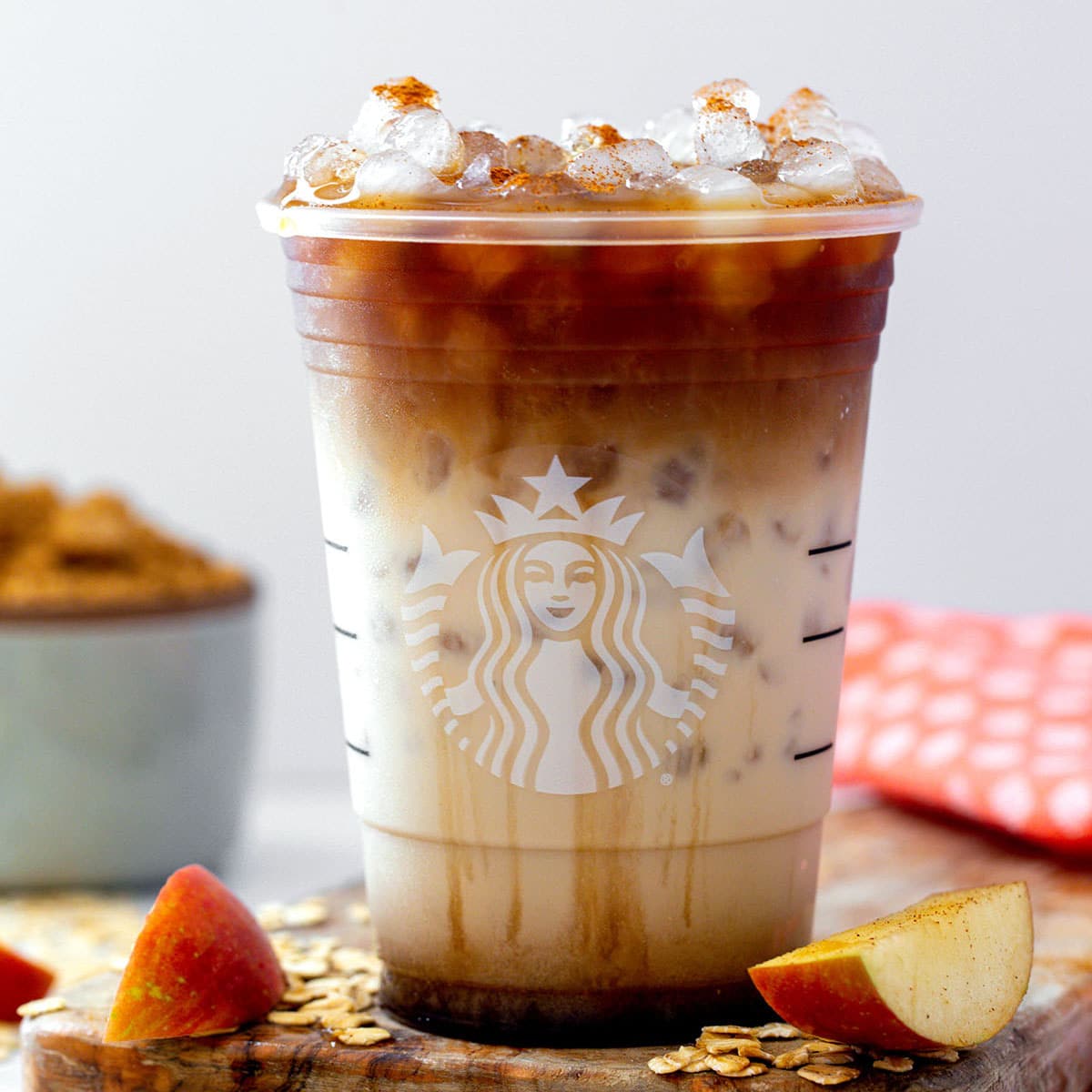 Closeup view of an Iced Apple Crisp Oatmilk Macchiato in a Starbucks cup with sliced apples and oats all around.