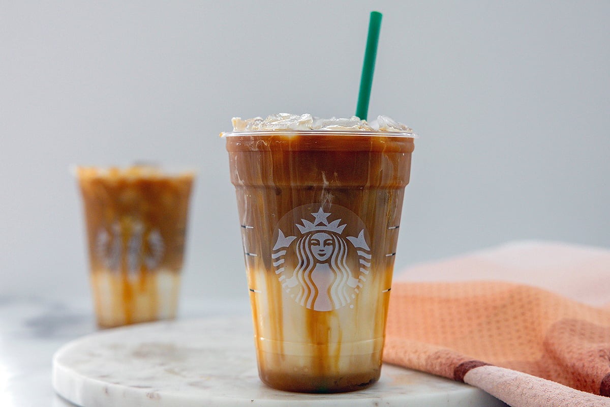 Landscape head-on view of an Iced Caramel Macchiato with green straw and second drink in background.