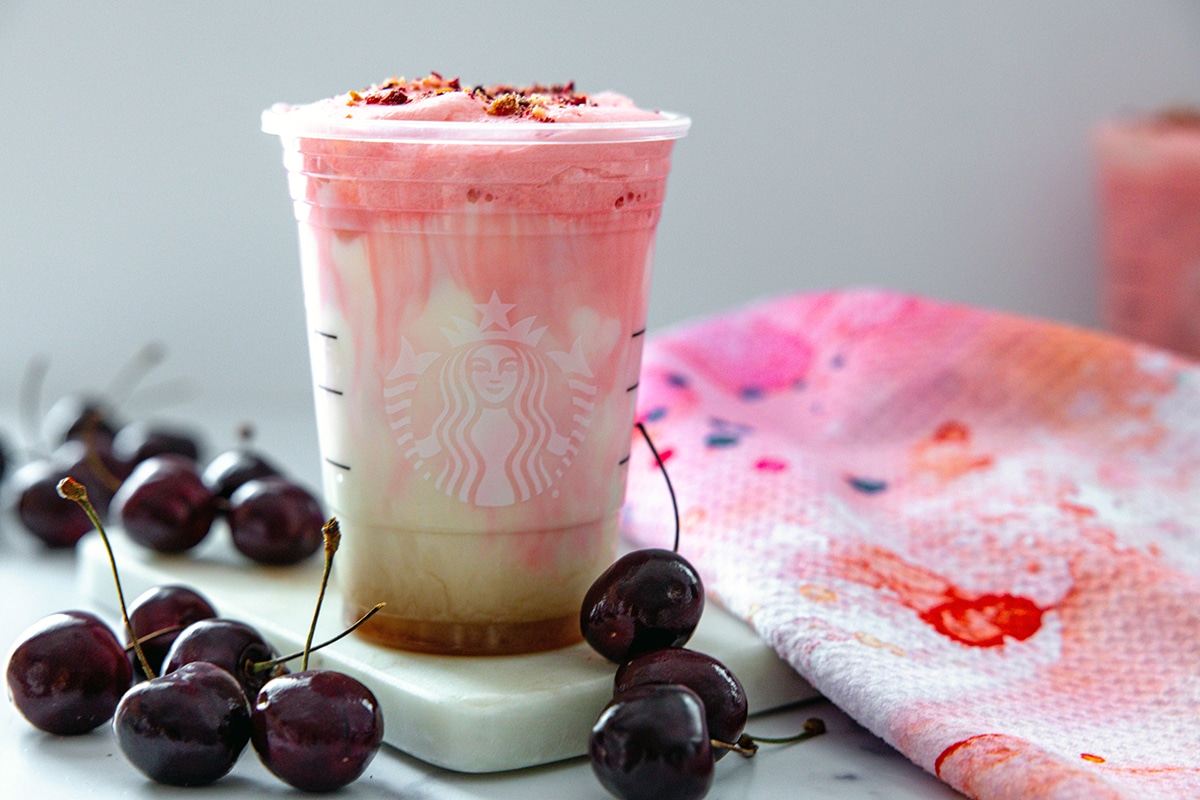 Landscape view of iced cherry chai latte drink with fresh cherries all around and pink, red, and white tea towel. 