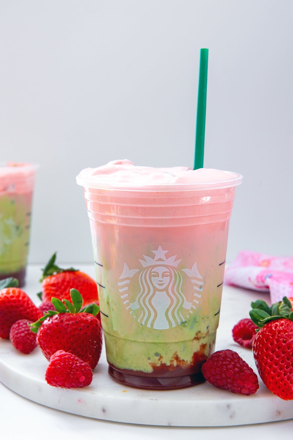 Head-on view of an Iced Double Berry Matcha with green straw with strawberries all around and second drink in background.