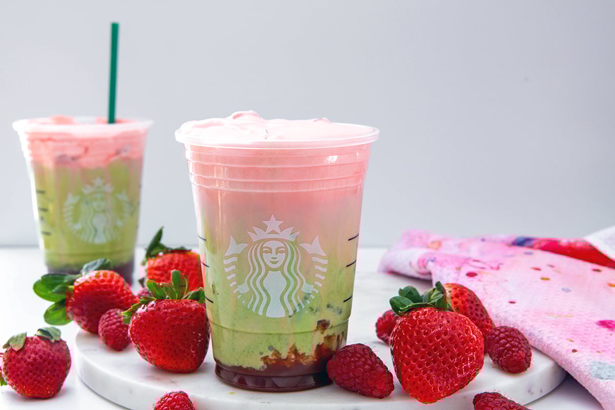 Landscape head-on view of two Starbucks cups with Double Berry Matcha Lattes in them with fresh strawberries all around.