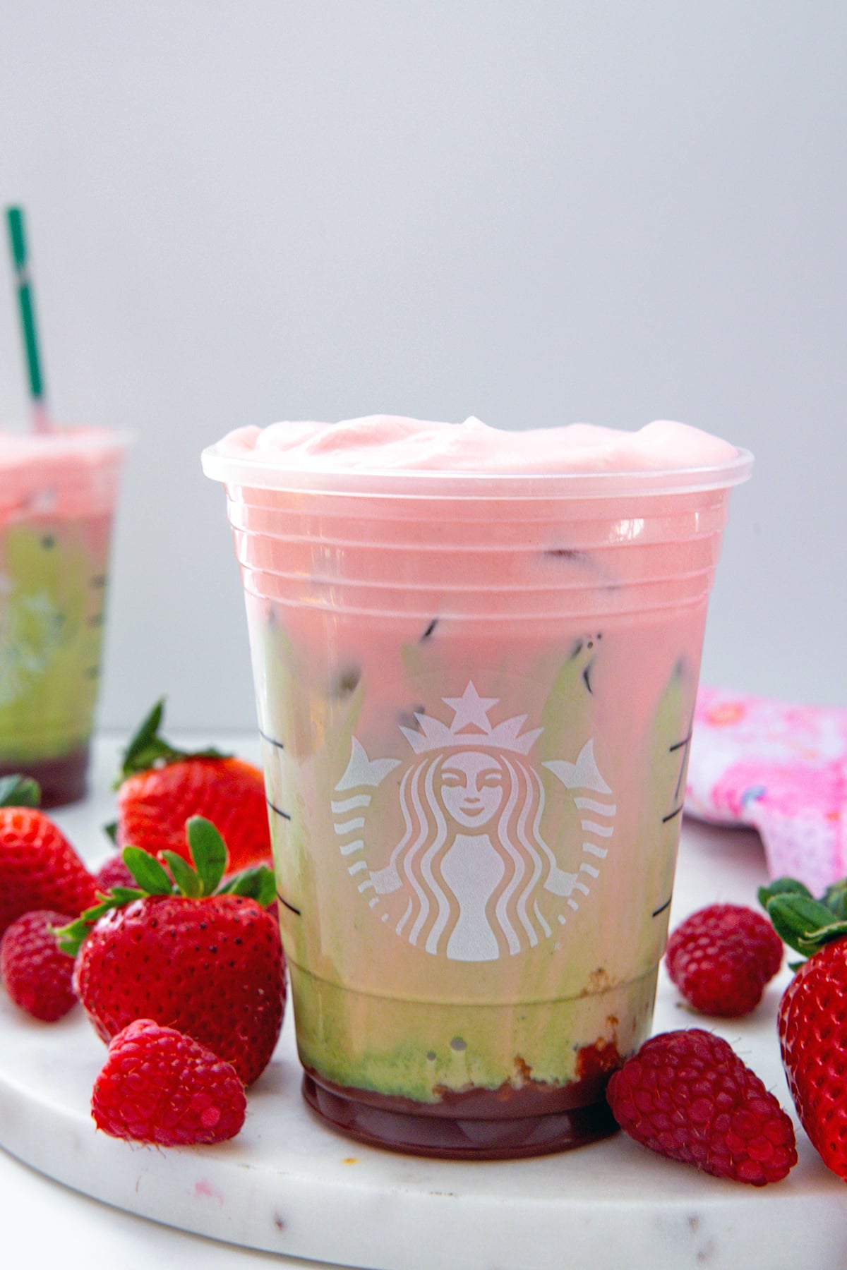 Head-on view of an Iced Double Berry Matcha in a Starbucks cup with raspberry cream cold foam blending into matcha and milk with strawberry puree on bottom of cup.