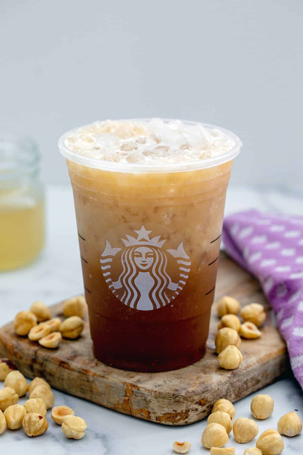 A Starbucks cup of Iced Hazelnut Oatmilk Shaken Espresso with hazelnuts all around and jar of hazelnut syrup in background.