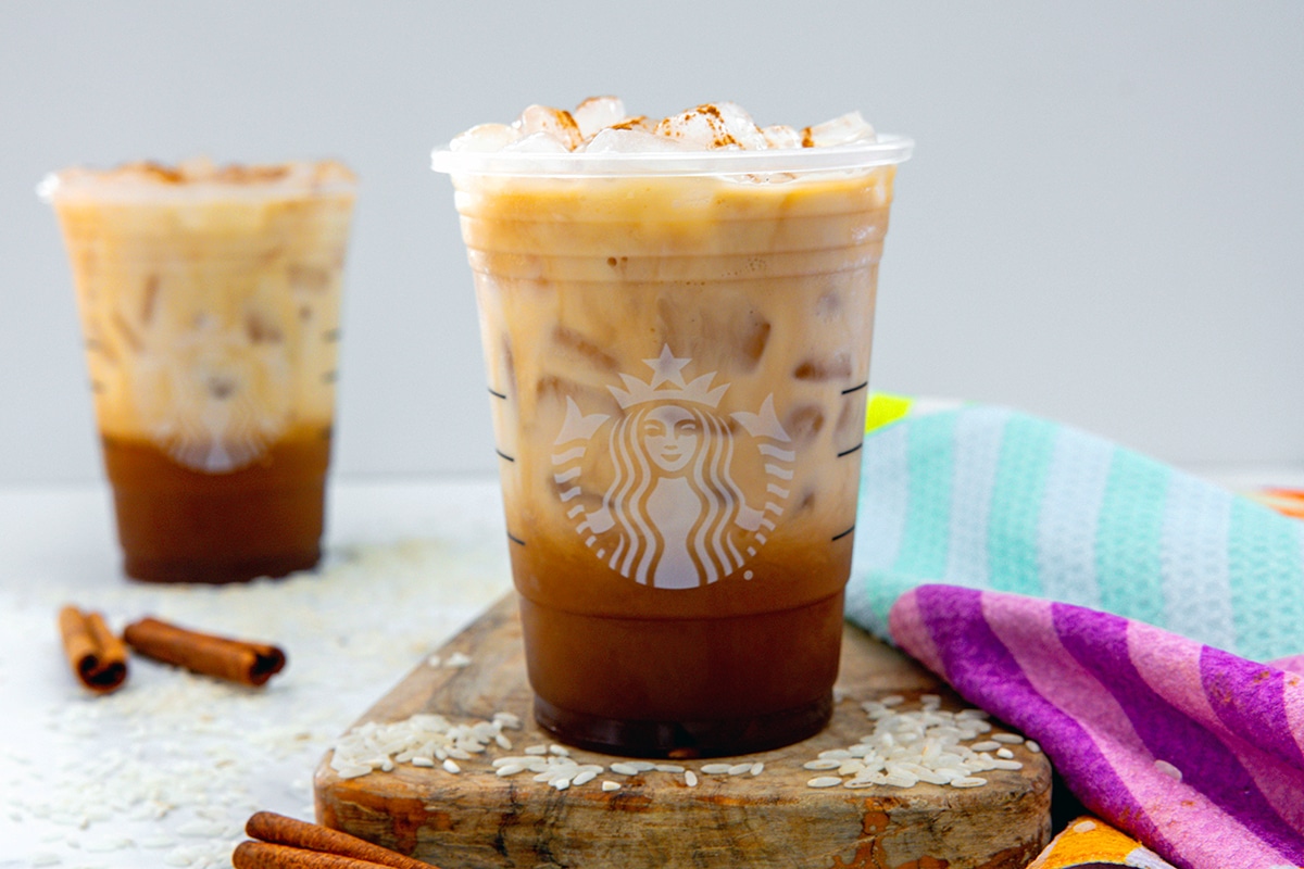 Landscape head-on view of a Starbucks Iced Horachata Oatmilk Shaken Espresso with second drink in backgrounf and rice and cinnamon sticks all around.