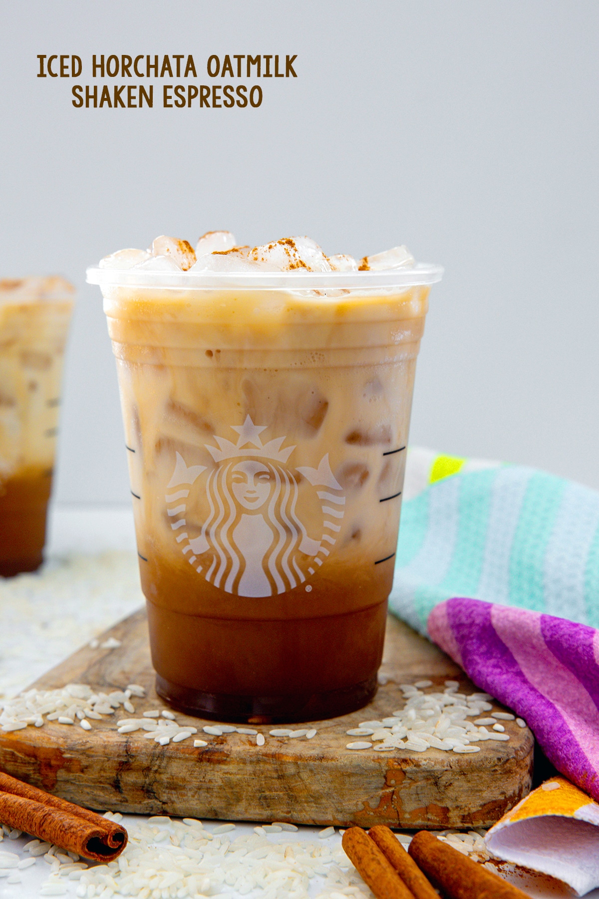 Head-on view of an Iced Horachata Oatmilk Shaken Espresso in Starbucks cup with white rice and cinnamon sticks all around and recipe title at top.