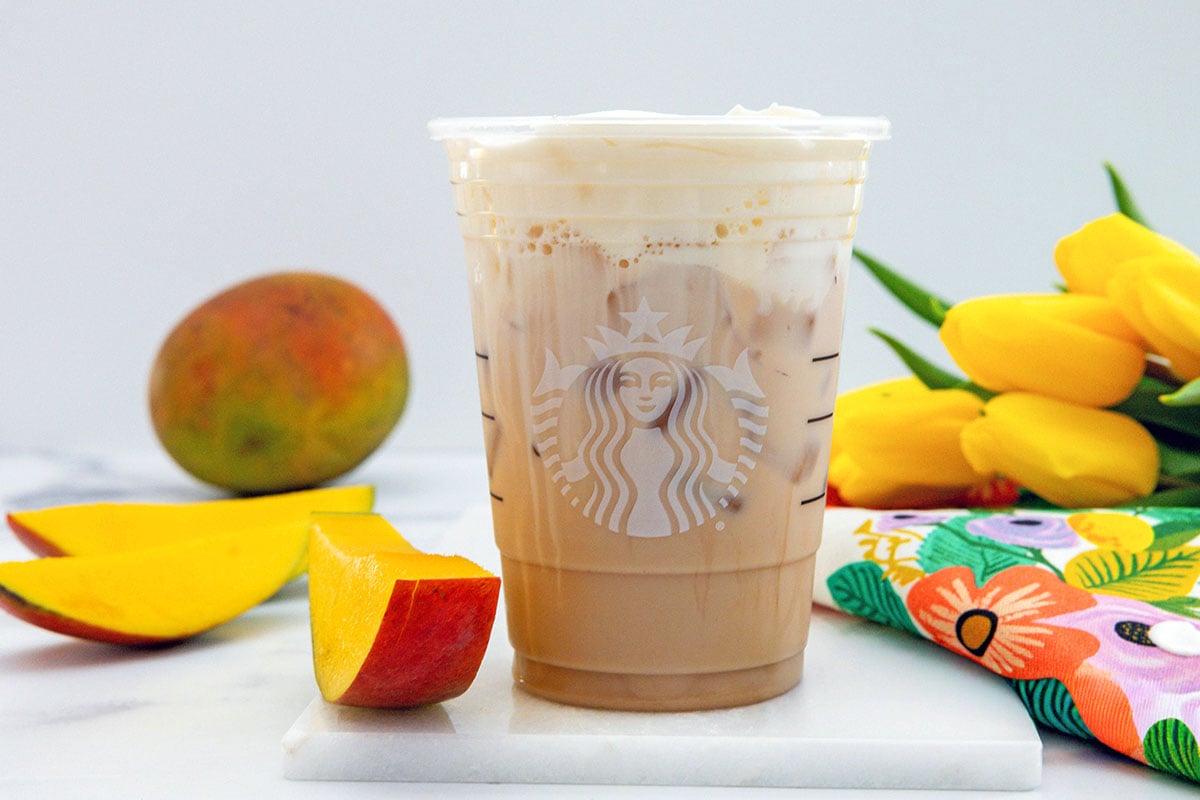Head-on landscape view of an Iced Mango Cream Chai in a Starbucks cup with sliced mangos all around and yellow tulips in background.