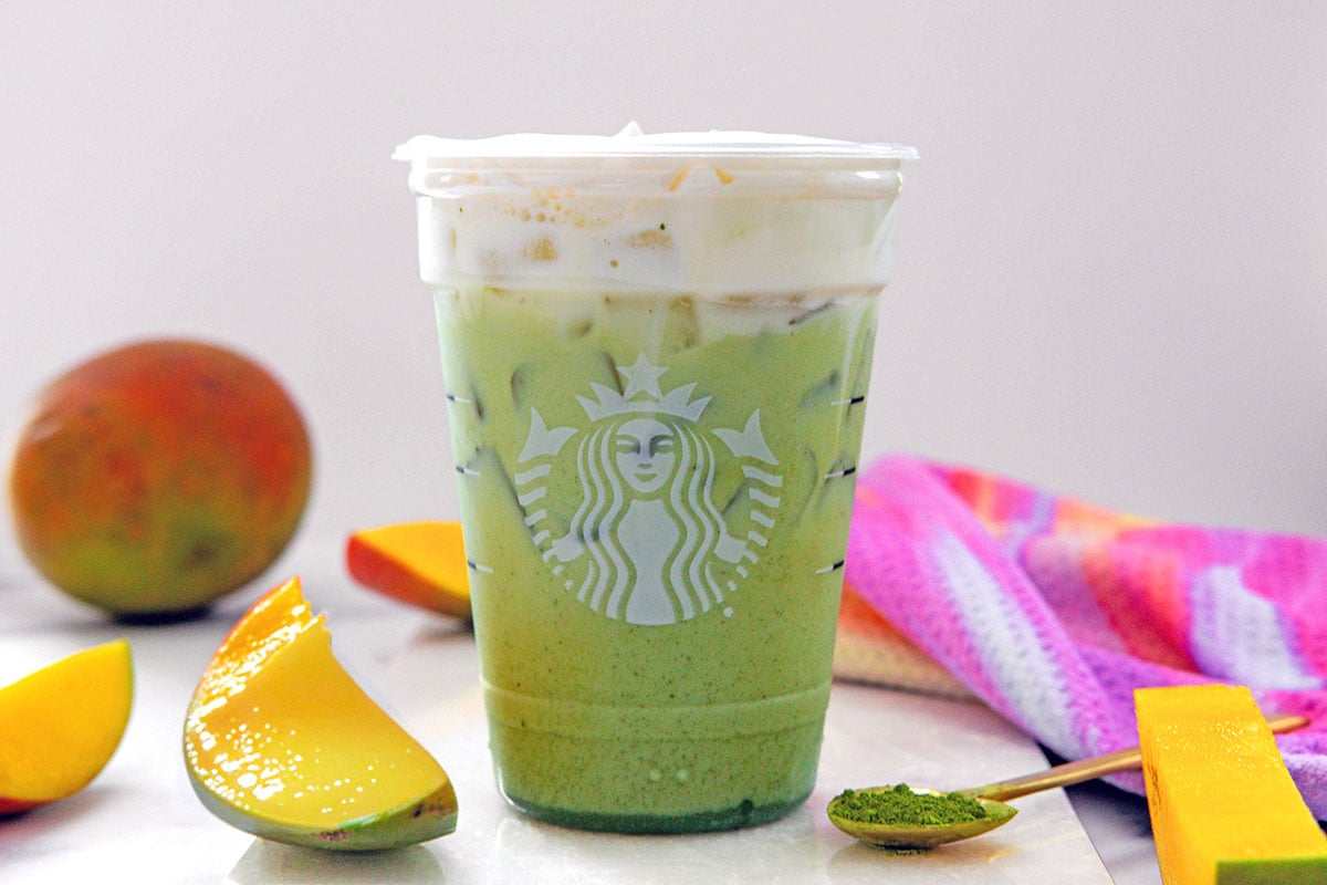 Head-on view of an Iced Mango Matcha in a Starbucks cup with sliced mango all around and whole mango in background.