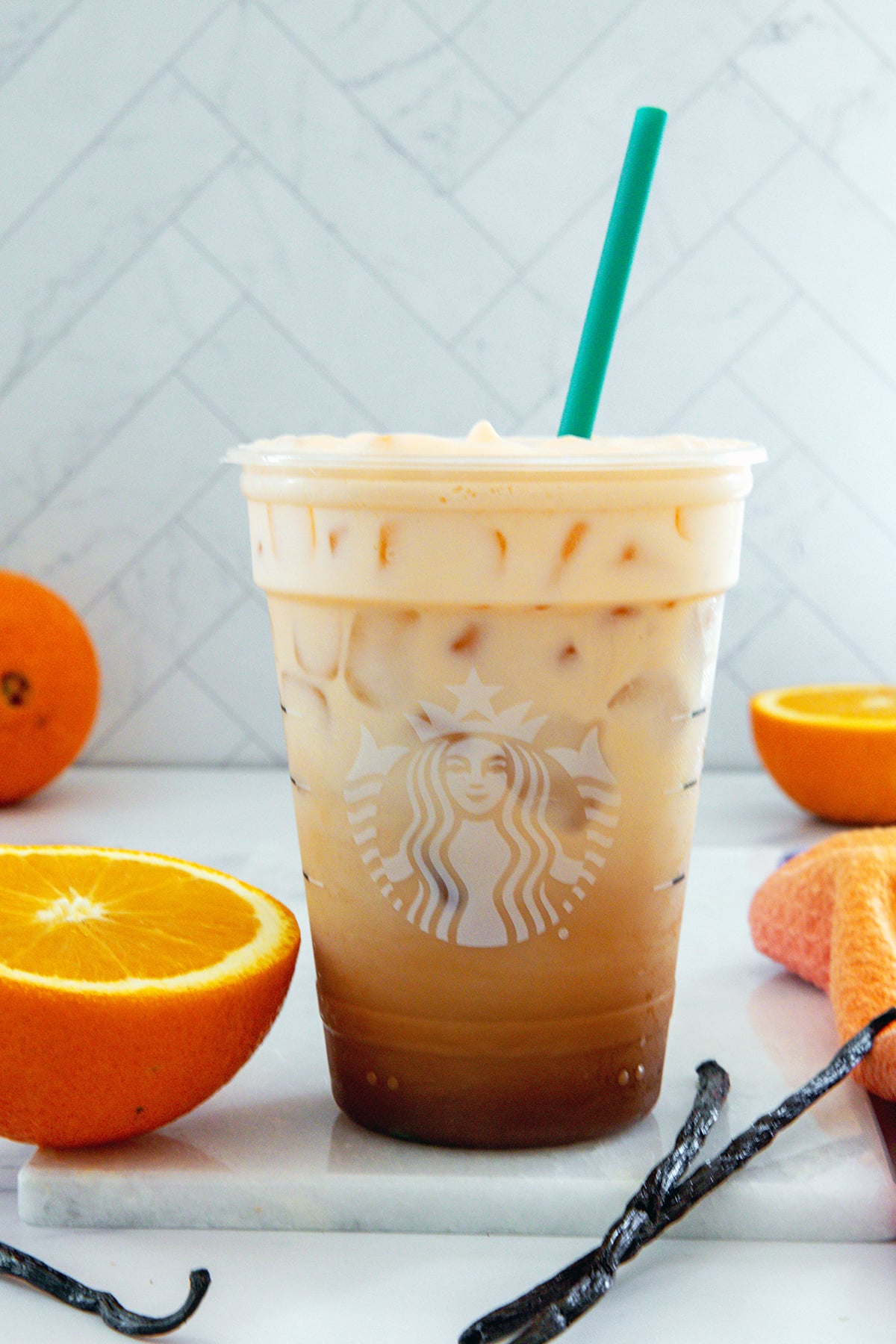 Head-on view of an Iced Orange Cream Latte in a Starbucks cup with oranges and vanilla beans all around.