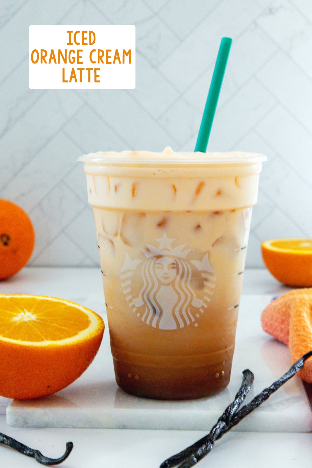 Head-on view of an Iced Orange Cream Latte in a Starbucks cup with oranges and vanilla beans all around and recipe title at top.