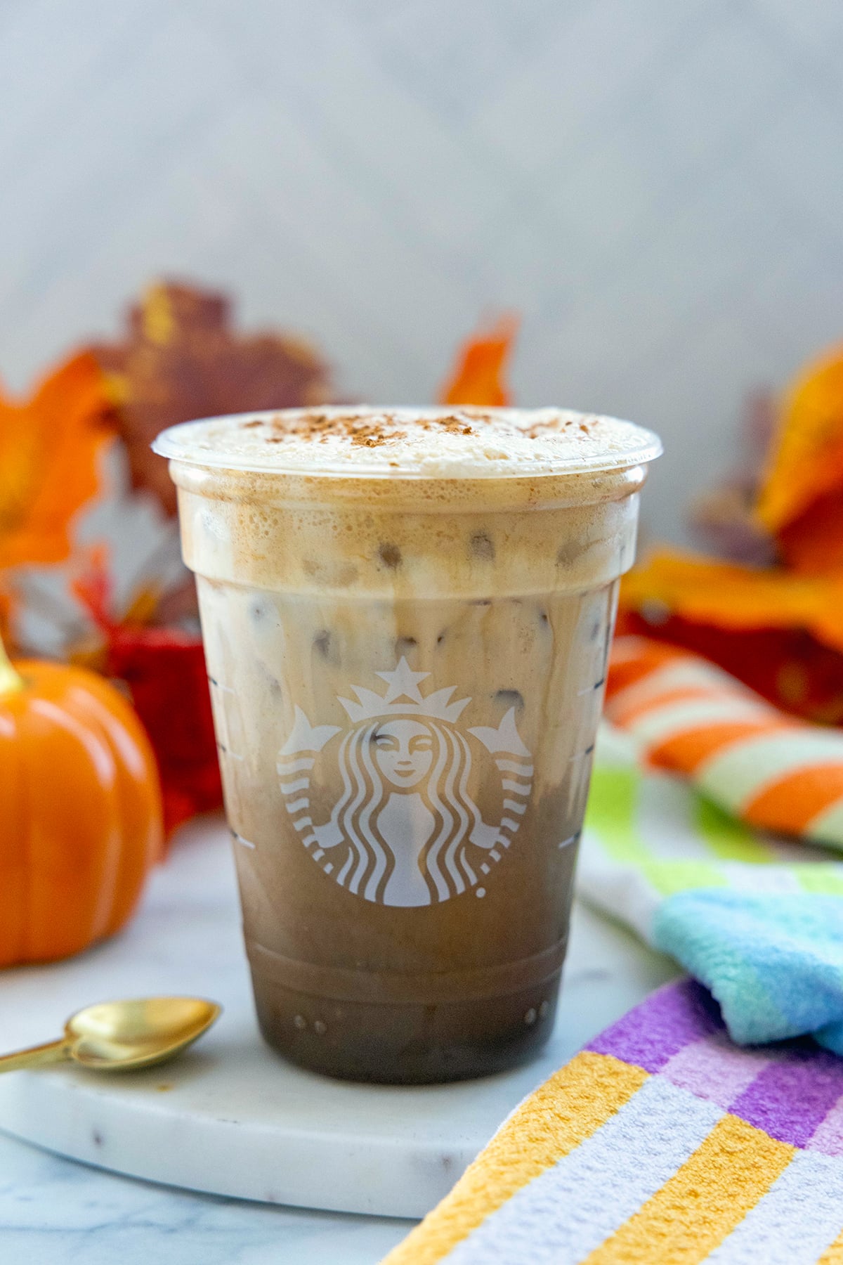 Head-on view of an Iced Pumpkin Cream Shaken Espresso in a Starbucks cup with pumpkin and fall leaves in background.