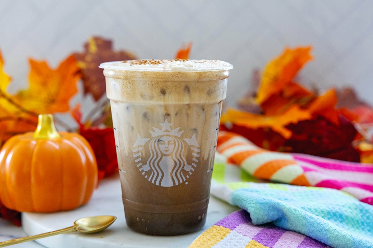 Landscape head-on view of Starbucks Iced Pumpkin Cream Shaken Espresso with spoon, pumpkin, and fall leaves in background.