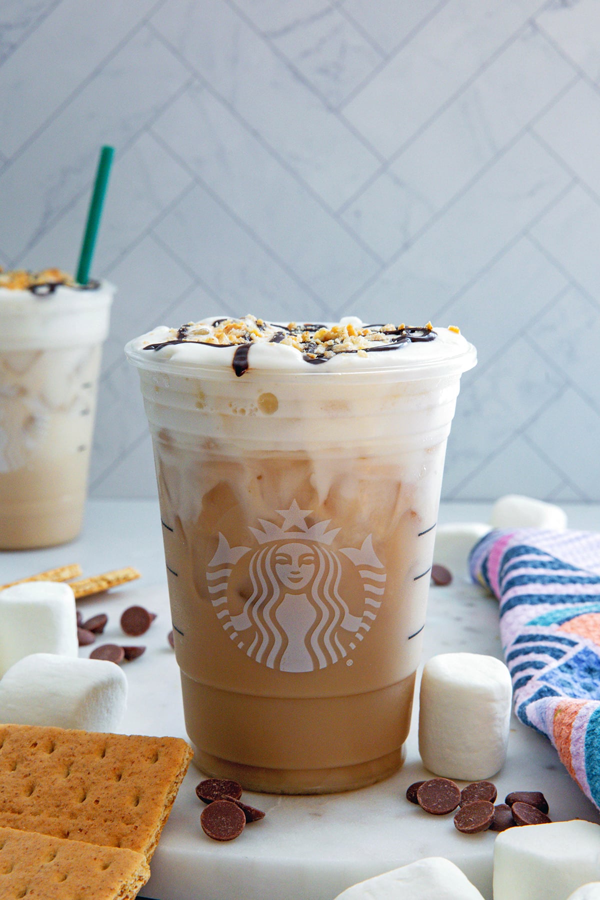 Head-on view of an Iced S'mores Chai in a Starbucks cup with graham crackers, chocolate, and marshmallows all around and second drink in background.