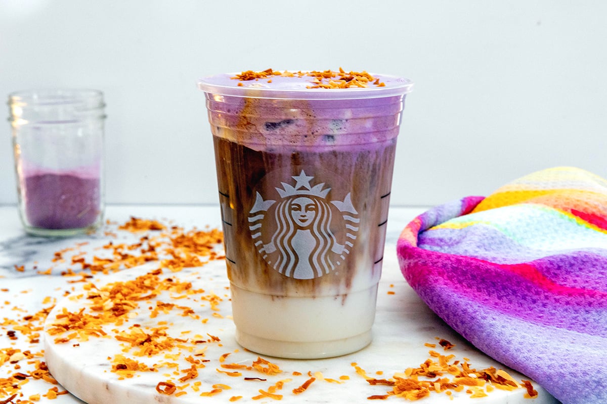Landscape head-on view of a Starbucks cup of Iced Ube Coconut Macchiato with toasted coconut all around and jar of ube powder in background.