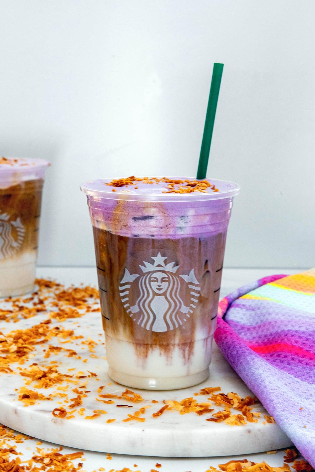 Head-on view of an Iced Ube Coconut Macchiato with green straw with second drink in background and toasted coconut all around.