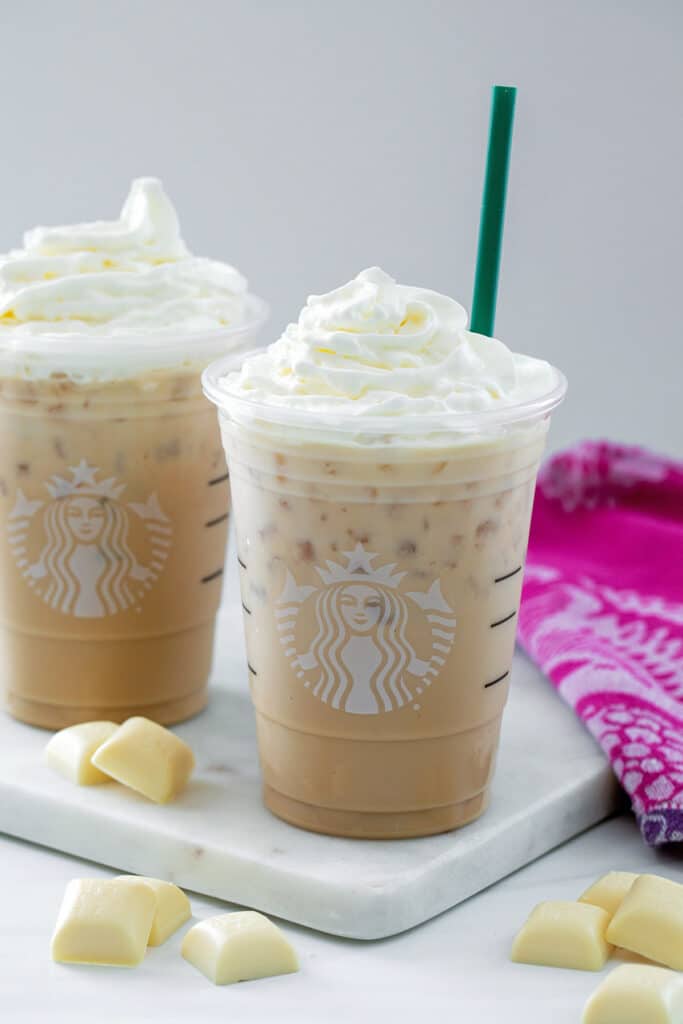 Two iced white chocolate mochas in cups with whipped cream with white chocolate squares all around.