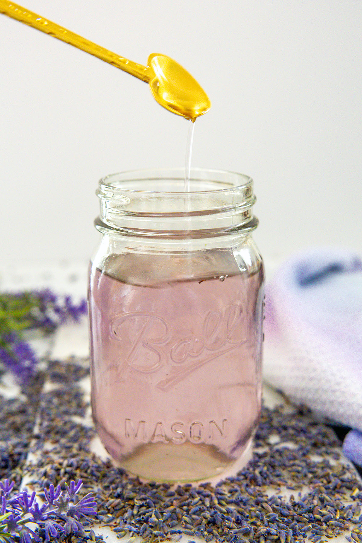 Head-on view of a mason jar lavender simple syrup with gold spoon drizzling into the jar.