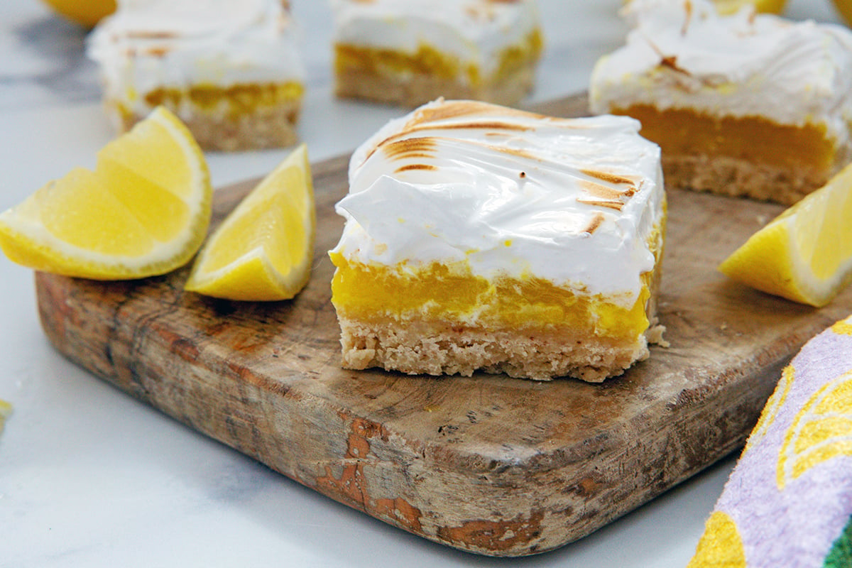 Landscape head-on view of a lemon meringue bar on a wooden board with lemon wedges.