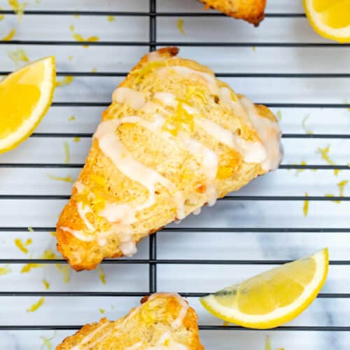 Lemon scone with icing drizzle on metal rack with lemon wedges.