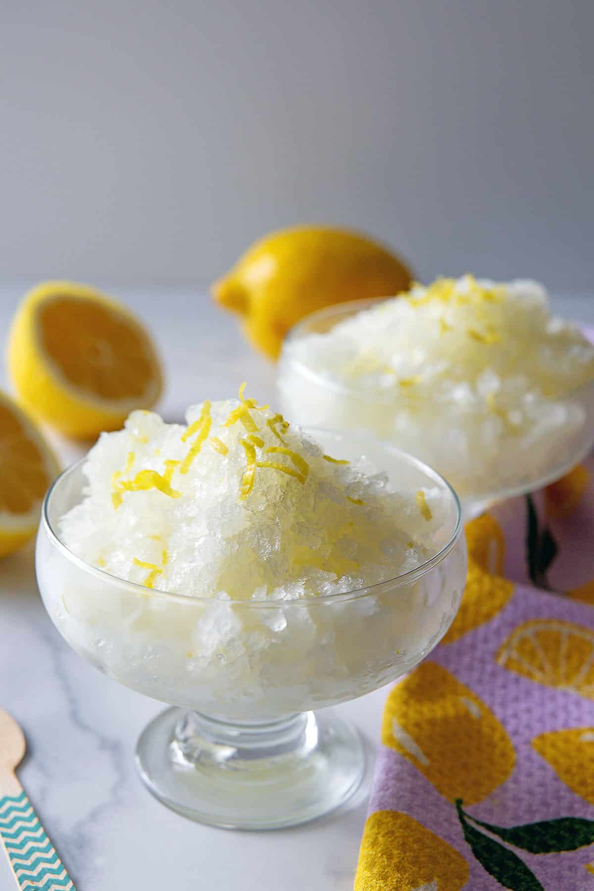 Two dishes of lemonade granita with lemon zest and fresh lemons in background.