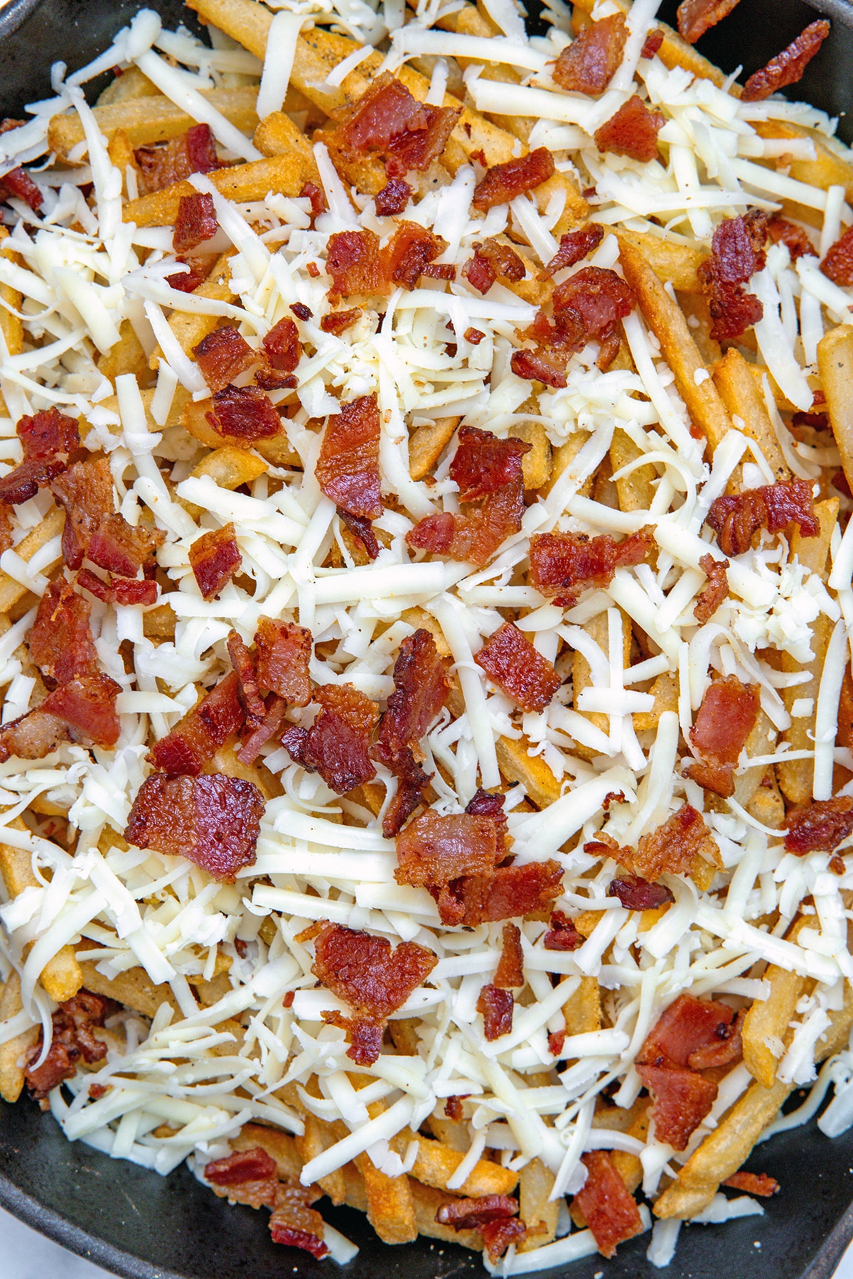 French fries in cast iron skillet topped with shredded cheese and crumbled bacon.