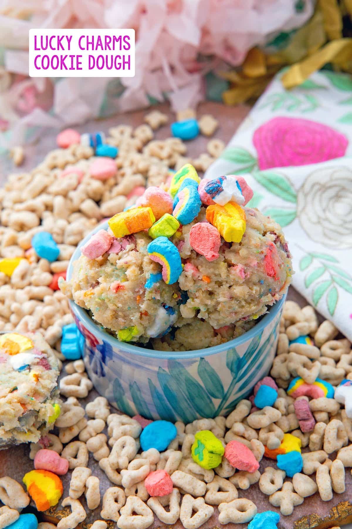 Overhead view of a bowl of Lucky Charms cookie dough with lots of colorful marshmallows and cereal all around with recipe title at top.