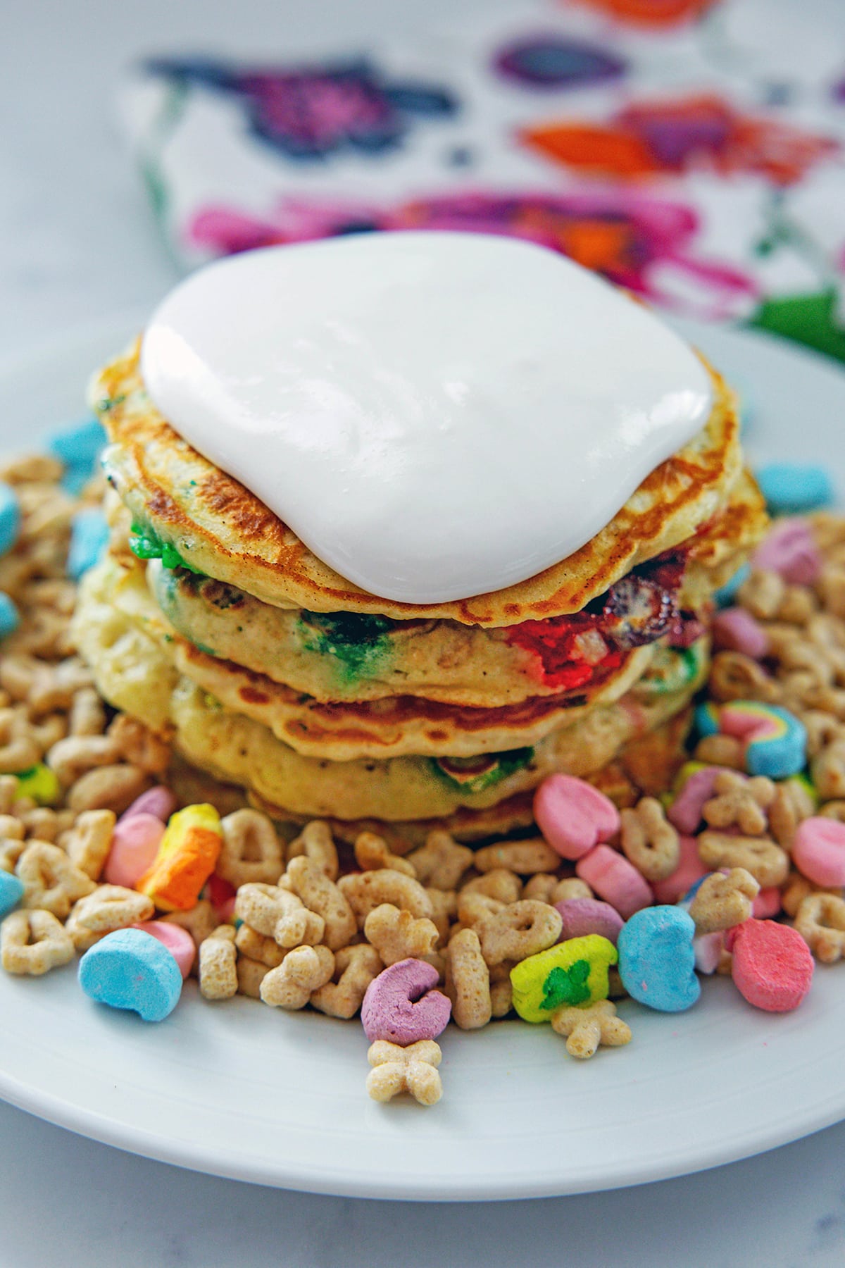 Stack of Lucky Charms pancakes topped with marshmallow sauce on a plate of cereal.