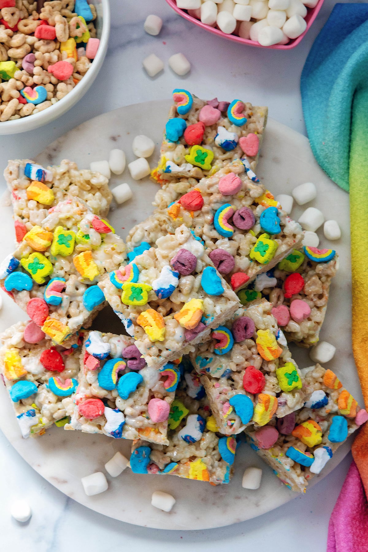 Overhead view of a platter of Lucky Charms Rice Krispies treats with mini marshmallows and Lucky Charms cereal all around.