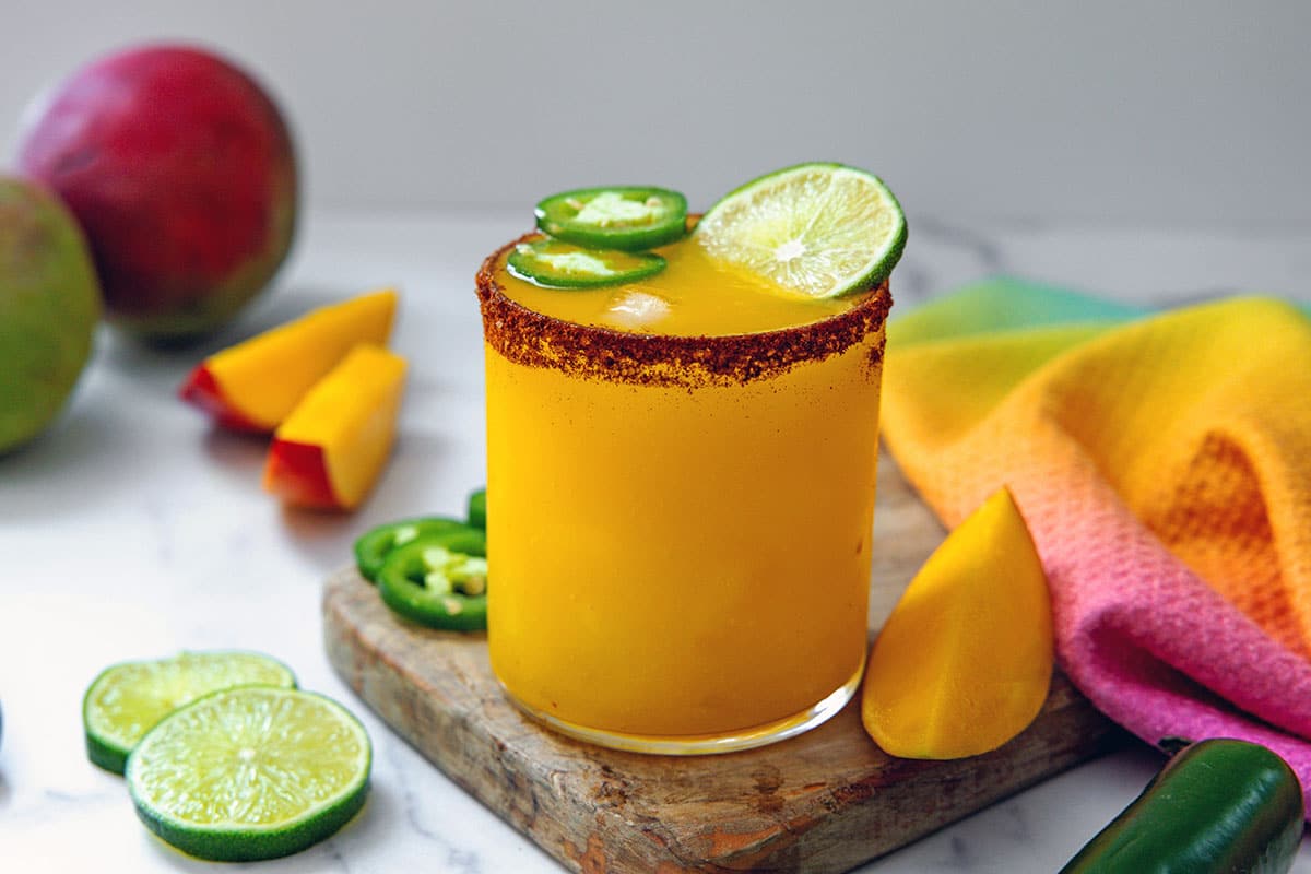 Head-on landscape view of a mango jalapeño margarita with sliced mango, limes, and jalapeños all around.