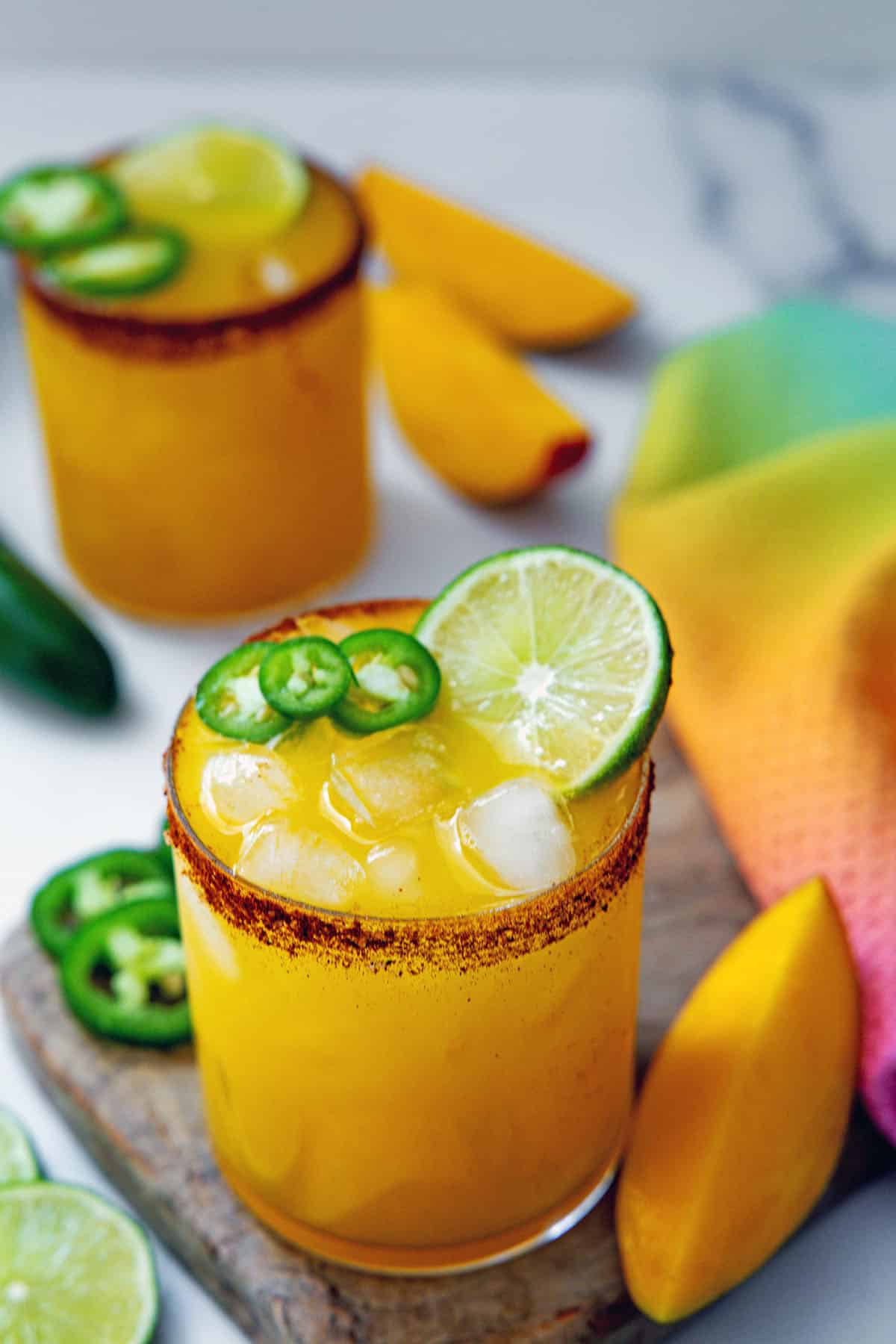 Two mango jalapeño margaritas with chili powder rims and sliced lime and jalapeño garnish with mango wedges all around.
