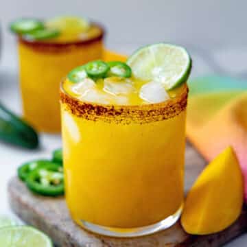 Closeup view of a mango jalapeño margarita with chili powder salt rim and sliced mangos, jalapeños, and limes in background.