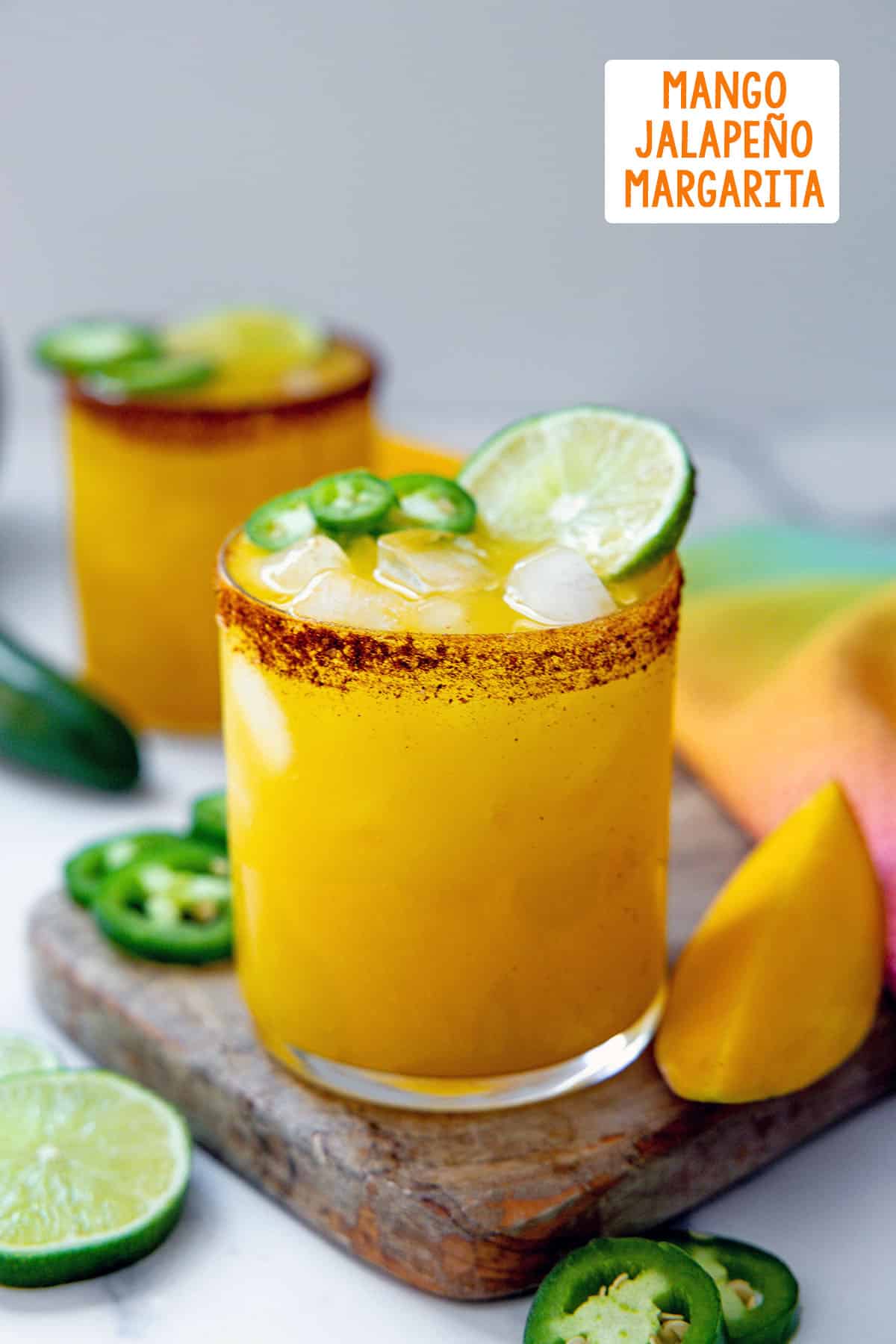 Head-on view of two mango jalapeño margaritas with chili salt rim with sliced limes and jalapeños all around and recipe title at top.