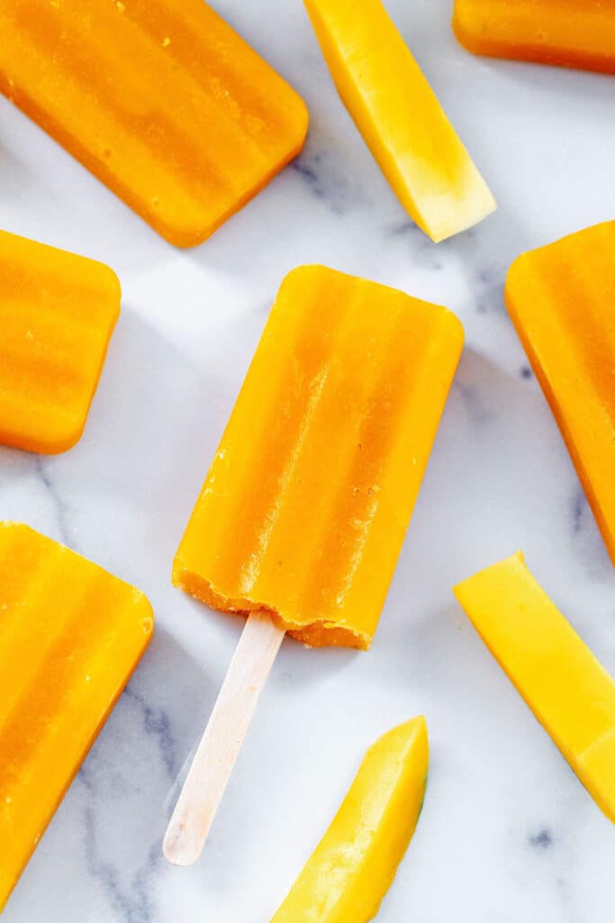 Mango popsicles and fresh mango slices on a marble surface.