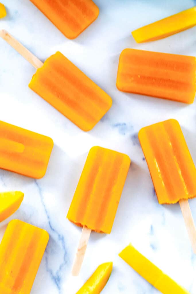 Multiple mango popsicles with mango slices on marble.