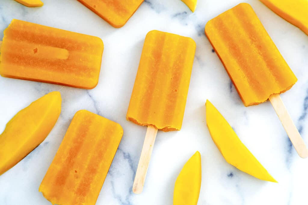 Landscape view of mango popsicles with fresh mango slices on marble table.