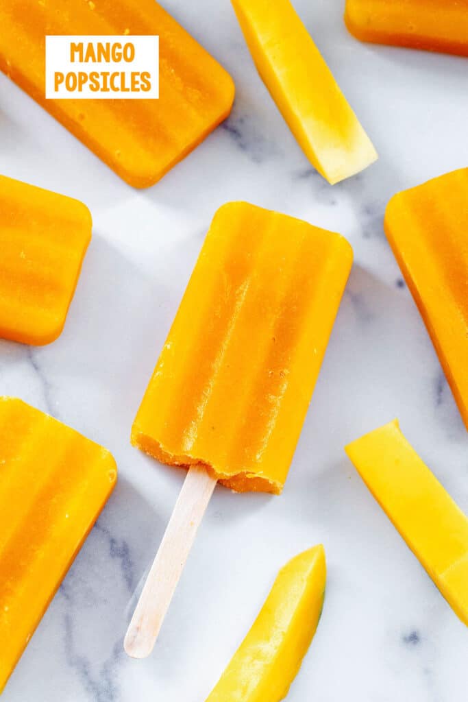 Overhead view of mango popsicles on a marble surface with mango slices all around and recipe title at top.