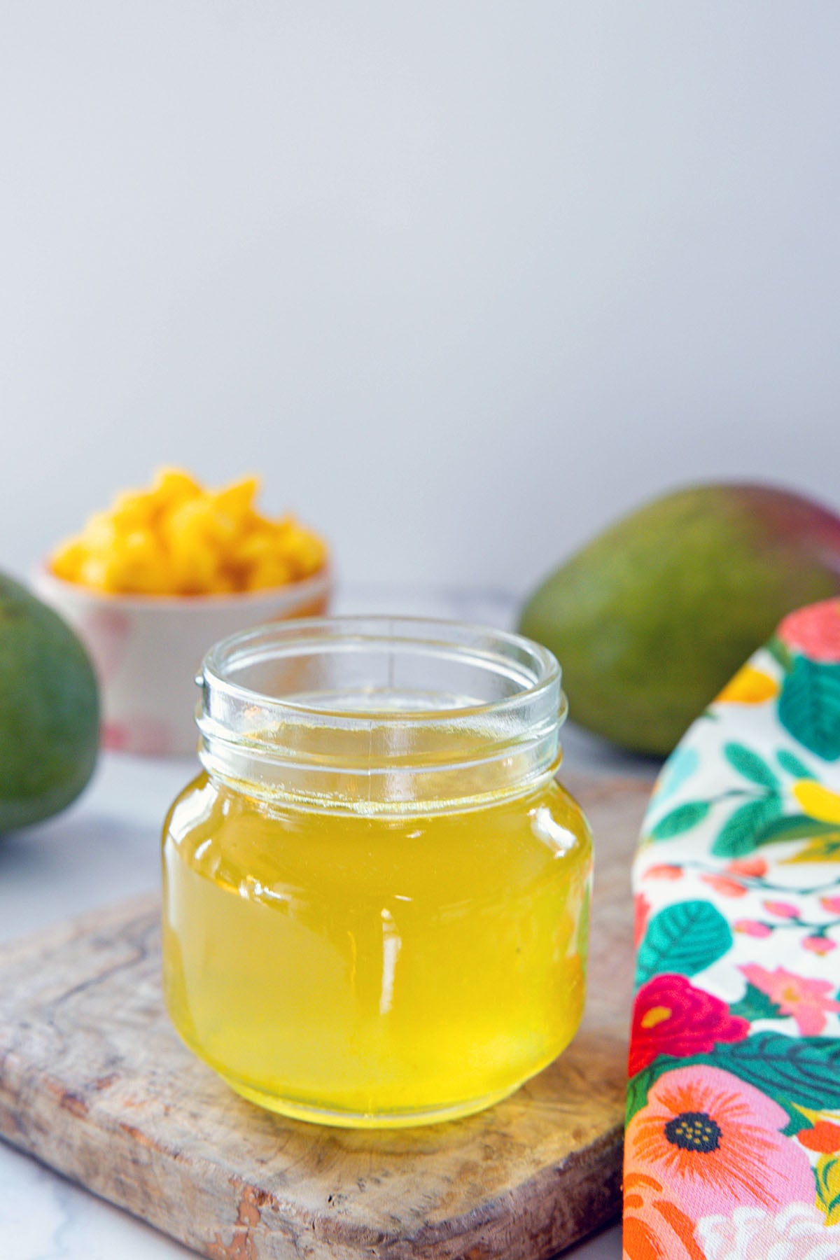 Head-on view of a small jar of mango syrup with mangos in the background.
