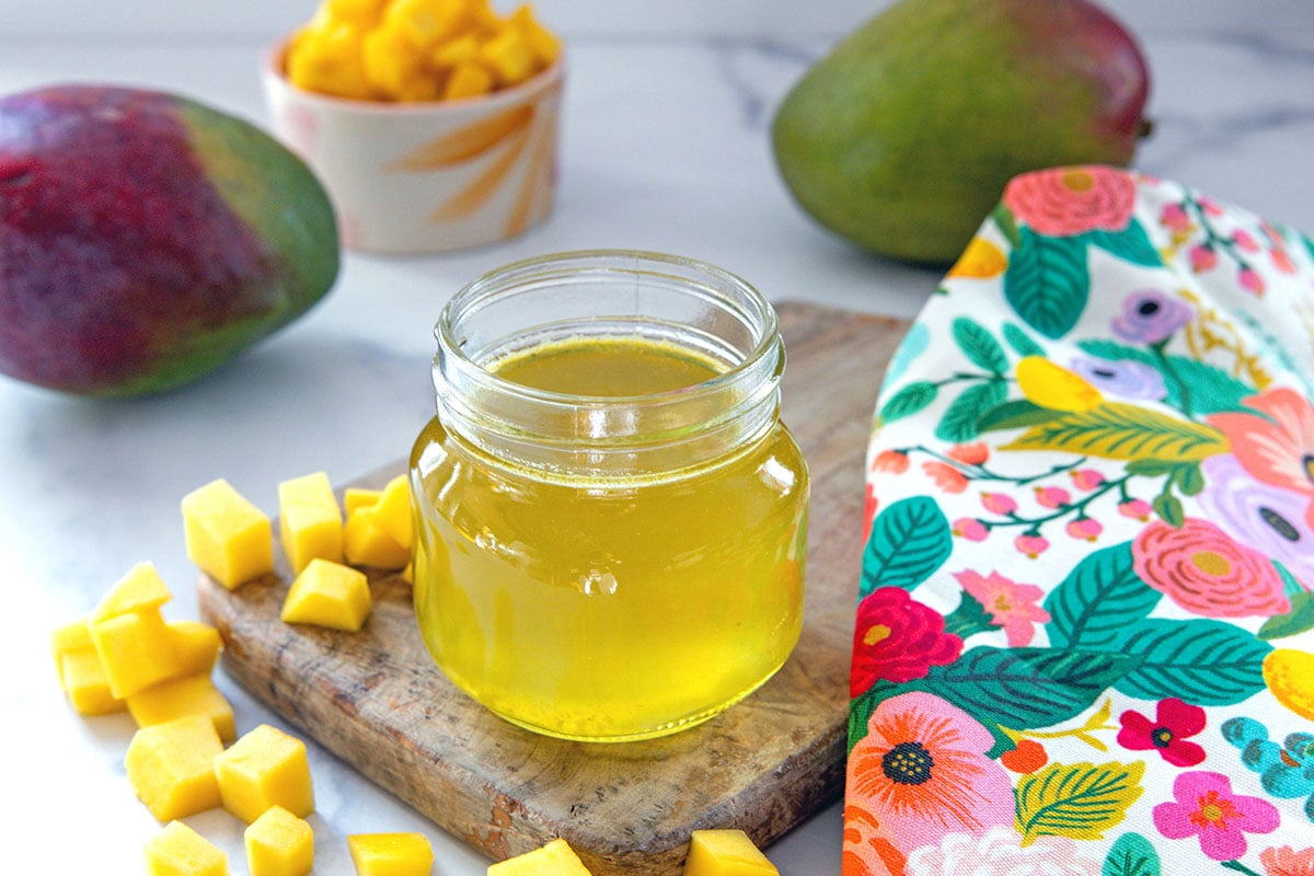 Landscape view of a small jar of mango syrup surrounded by chopped mango and whole mangoes.