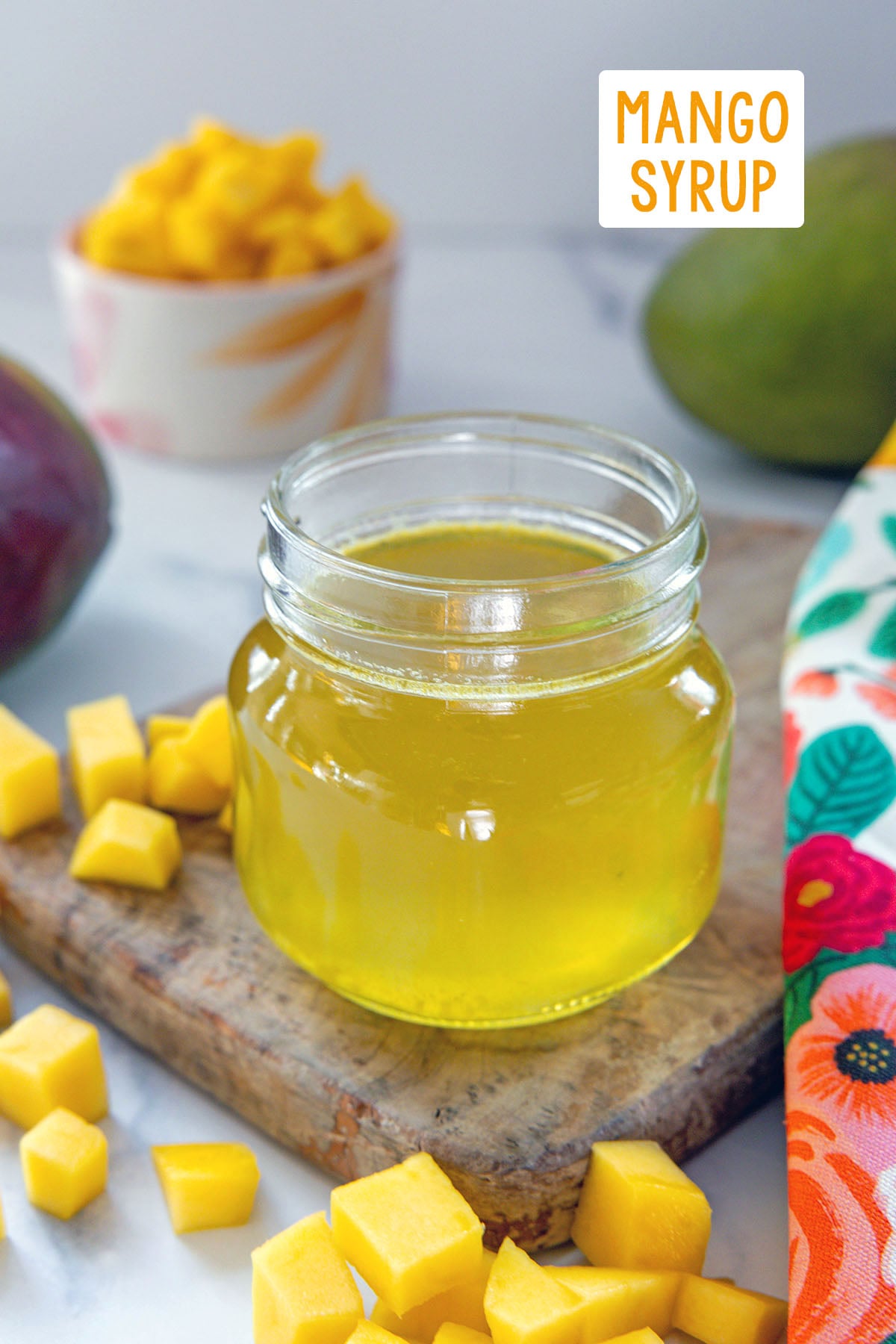 Small jar of yellow mango syrup with chopped mango all around and recipe title at top.